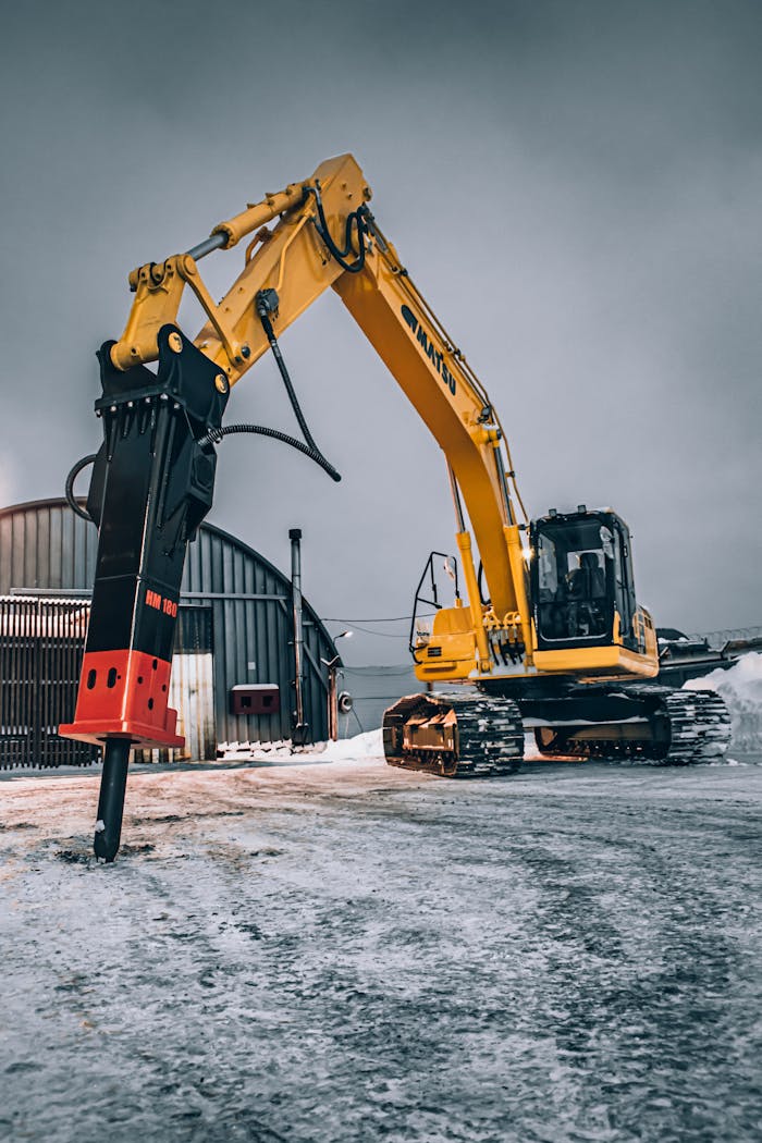 gallery-1 A yellow excavator with a hydraulic breaker parked on a snowy construction site.